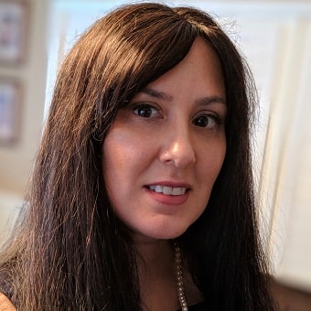 A woman with long brown hair, wearing a black top and a pearl necklace, smiles slightly at the camera indoors, her serene demeanor suggesting the positive effects of psychotherapy.