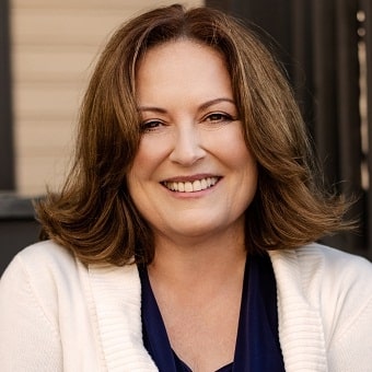 A person with shoulder-length brown hair smiles, wearing a white cardigan over a dark top, embodying the warmth often found in psychotherapy sessions.