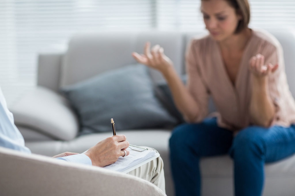 A woman gestures with her hands while sitting on a couch, engaging in a psychotherapy session with one of the best doctors, who takes notes attentively.