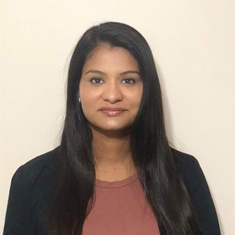 Portrait of a person with long dark hair, wearing a black blazer and a maroon top, standing against a plain light background, embodying the serene professionalism often found in clinicians online.