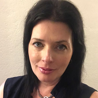 A person with long dark hair and a neutral expression, wearing a black top and necklace, stands in front of a plain light-colored wall, embodying the calm professionalism you'd expect from the best doctors in group counseling sessions.