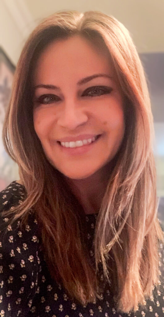 A person with long, straight hair and a floral patterned top smiles at the camera in a well-lit setting, exuding warmth reminiscent of group counseling sessions.