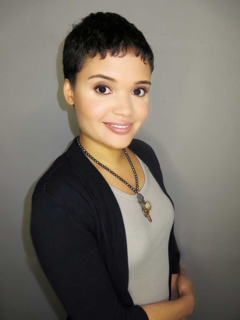 A person with short black hair and a dark cardigan smiles at the camera, exuding warmth akin to a welcoming psychotherapist in a mental health clinic. Against the gray background, their necklace with two pendants adds a touch of personal style.