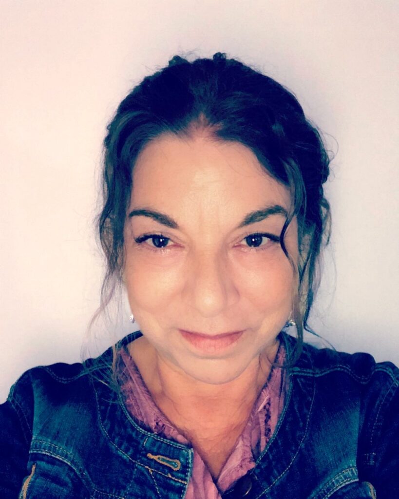 A woman with dark hair and a denim jacket smiles subtly at the camera against a plain background, exuding the warmth of someone who believes in the power of psychotherapy.