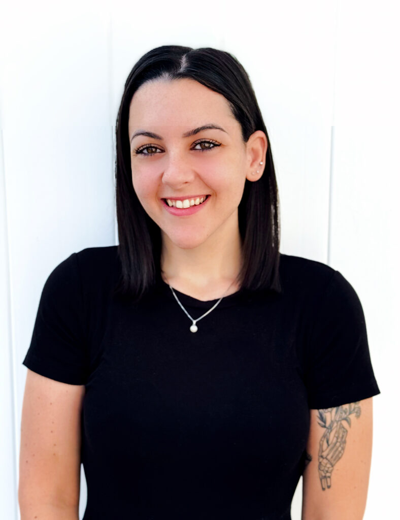 A person with straight black hair, wearing a black shirt and a necklace, smiles while standing against a white background. They have a tattoo on their left arm, perhaps reflecting their journey at a mental health clinic.