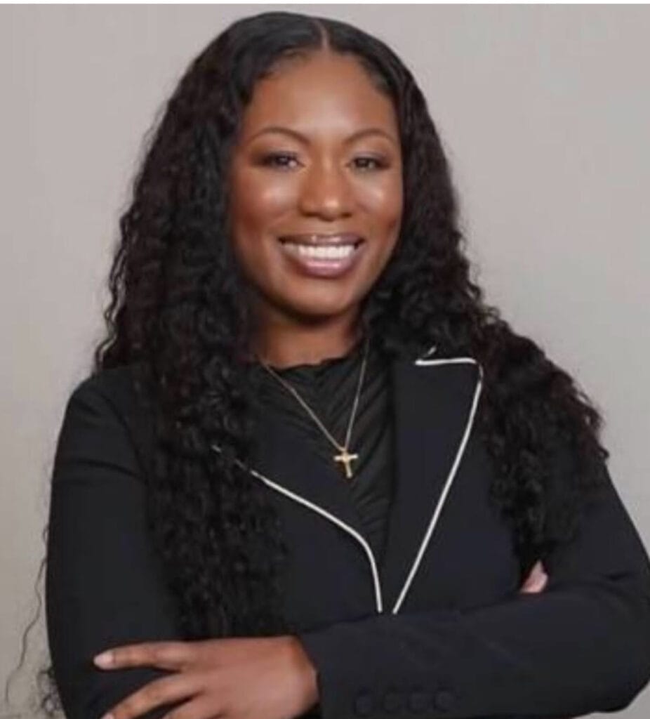 A woman with long curly hair is smiling, wearing a black blazer with white trim and a small cross necklace, exuding warmth and professionalism typical of a mental health clinic setting.