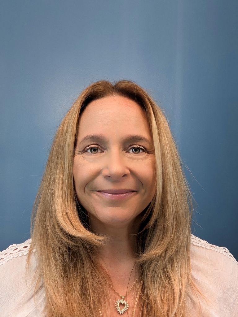 A person with long, blonde hair stands against a blue background, wearing a white top and heart-shaped necklace, smiling at the camera—capturing the warmth often found in a mental health clinic setting.