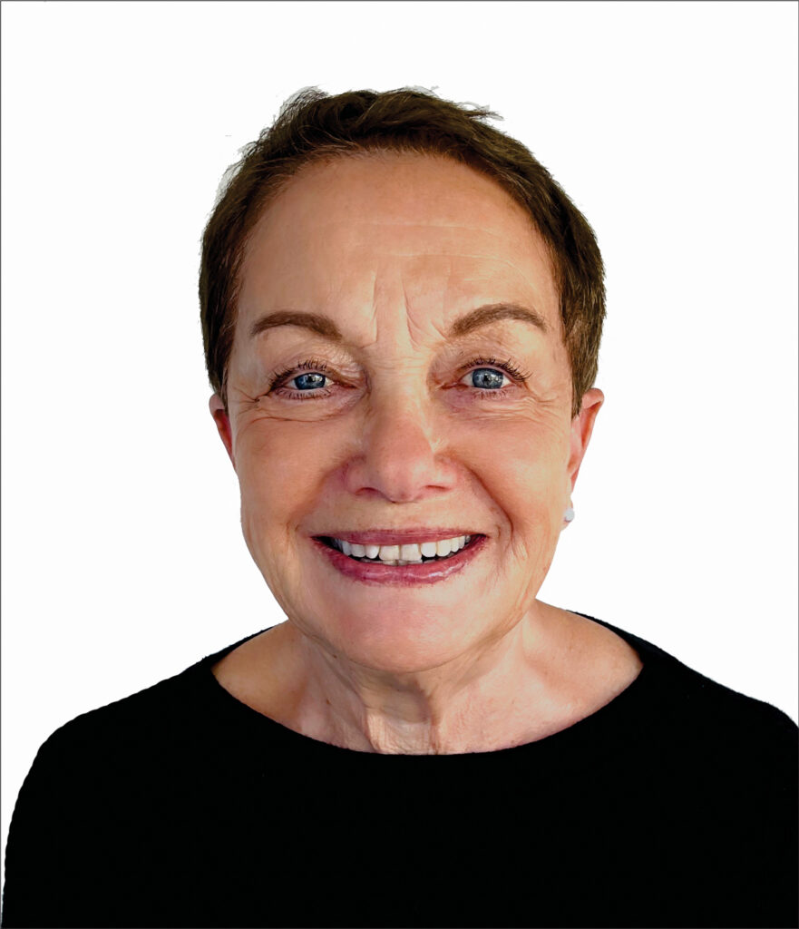 A smiling elderly woman with short hair, perhaps a dedicated practitioner engaged in online psychotherapy, is wearing a black top against a plain white background.