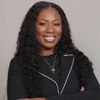 A smiling woman with long, curly hair wears a black blazer and a necklace with a cross pendant, standing against a neutral background, ready for her telepsychiatry session.