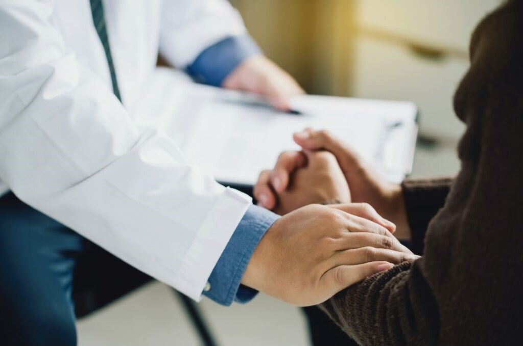 A doctor in a white coat holds a clipboard while providing psychotherapy, comforting the patient by placing a hand on their arm.