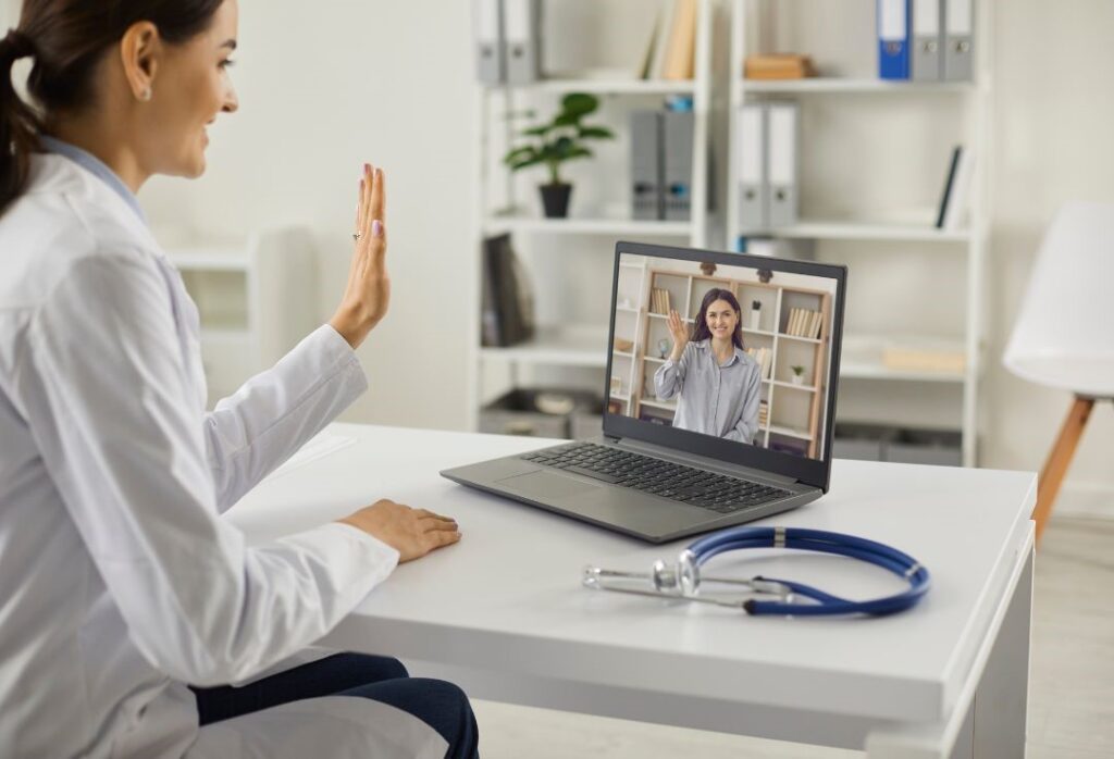 A doctor in a white coat waves during a video call from a mental health clinic, connecting with a patient on the laptop. A stethoscope is on the table, signaling the dedication of one of the best doctors committed to comprehensive care.