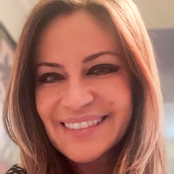 A smiling person with long brown hair, wearing makeup, sits in a softly lit indoor setting, possibly reflecting on the positive impact of group counseling.